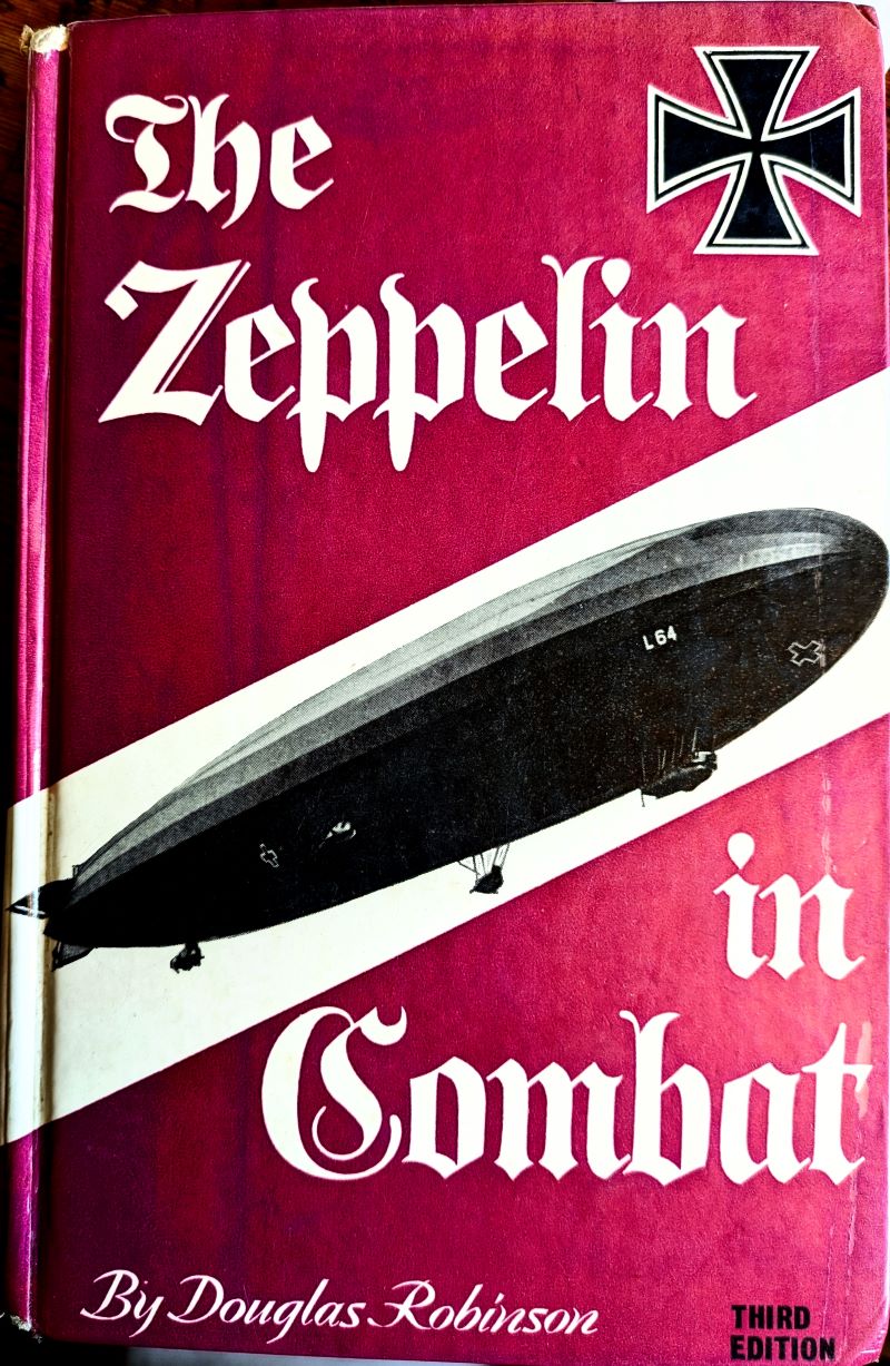 Picture of book cover showing a Zeppelin against a white background across an otherwise pink cover with German Iron Cross medal shown in top right corner and with title and author in white text .